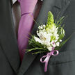 This image shows a finely dressed person ready for a special event in Epping, captured from the chest up so the focus rests on their lapel and tie. The suit jacket is a deep charcoal grey with a smooth, slightly matte finish, its lapel pressed with exacting care, hinting at bespoke tailoring that wouldn't look out of place at an Epping church wedding or a celebration near Theydon Bois. Beneath the jacket, a light dress shirt with an ultra-fine dark check offers a subtle pattern, while a solid lavender necktie runs straight and smooth, its satin sheen reflecting gentle highlights in the surrounding light. Pinned to the left lapel is a meticulously crafted boutonniere that feels both fresh and romantic. Several white Peruvian lilies are the stars of the piece, each blossom opening to reveal slender petals painted with delicate reddish-brown streaks and soft hints of violet, leading into yellow-toned throats with visible stamens. Standing tall at the centre is a conical green bud reminiscent of tuberose, adding structure, height, and a cool, springlike green tone. Around these main elements, tiny white clusters of gypsophila create a cloud-like softness, while a mix of greenery-fine, spiky blades, feathery fronds, and broader deep green leaves-adds texture and depth, much like the layered foliage in Epping Forest. The stems are gathered and wrapped with a satin ribbon in a soft lilac shade, tied into a precise little bow that mirrors the lavender tie. Together, the flowers and formalwear express quiet confidence and heartfelt anticipation, ideal for weddings, proms, or milestone events across the Epping community.
