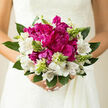 This image shows a romantic bridal bouquet held close to the waist of a bride, the creamy fabric of her dress providing a soft backdrop that lets every petal stand out clearly. At its heart are generous magenta peonies, rounded and luxurious, with petals that seem to tumble over one another in a swirl of raspberry and plum hues, like the richest blooms you might find in a sheltered Epping back-garden. Surrounding them, white alstroemeria form a delicate ring, their petals slightly flared and gently curled at the tips, each one etched with fine burgundy and amber streaks that draw the eye inward and add an almost lace-like texture. Sprinkled among these are pale green buds carried on slender stems, just beginning to open, suggesting growth, renewal, and the start of something beautiful, much like the fresh shoots that line the edges of the roads leading out from Epping towards the countryside. Deep and mid-green foliage anchors the arrangement, some leaves broad and glossy, others more slender and feathery, together creating a cool, leafy cradle that sets off the bright blooms. The natural daylight in the photograph falls softly from one side, giving a gentle sheen to the peony petals and illuminating the intricate markings on the alstroemeria, while subtle shadows along the stems add structure and depth. The bouquet gives the impression of a light, green, slightly sweet fragrance, echoing a morning walk past the gardens off Epping High Street. Everything about the composition feels poised between classic and contemporary, an elegant, thoughtfully crafted design perfect for wedding photography or cherished memories of a special day in Epping.