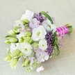 Intricately arranged by an Epping florist, this hand-tied bridal bouquet radiates springtime romance atop a pale, softly textured linen reminiscent of local artisan tables. Creamy-white Lisianthus, their ruffled forms open and welcoming, cluster centrally as the heart of the bouquet, surrounded by pastel lavender and dusky purple accents. Dainty, tightly closed lisianthus buds and lime-tinted green sprigs weave through the design, infusing it with freshness and a sense of growth-a visual salute to the lush landscapes around Epping Forest. Energetic star-shaped purple allium punctuate the bouquet, their bold, spiky forms adding depth and modern edge to the otherwise classic palette. Sinuous blades of vivid green foliage loop and arc gracefully, framing the arrangement with gentle movement. The hand-gathered stems are bound in a vivid magenta satin ribbon, finished with a draping of gleaming white pearls-a stylish flourish that speaks to the joy and celebration of a wedding morning. Filtered daylight sets the colors aglow, casting buttery and pastel notes that feel as cool and refreshing as a dawn breeze over Epping fields. This bouquet is designed to be cherished, symbolising new beginnings, friendship, and the artistry rooted in the heart of Epping's close-knit community, perfect for wedding features or local bridal inspiration.