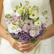 This bouquet appears as a piece of living poetry held close to the chest, the kind of bridal design you might see carried down the aisle in an Epping church just off the High Street. Soft, unfolding whites and delicate pastel lilacs are the main characters here, with lisianthus and stock intertwining in gentle waves of texture. The lisianthus blossoms are creamy and smooth, their petals curving like silk ribbons, while the pastel stocks rise in clustered spires, each floret slightly ruffled as if brushed by a faint breeze drifting over Epping Forest. At the base, a distinct halo of lavender allium forms a textural crown: hundreds of tiny, star-shaped florets create a cloud of muted purple that grounds the bouquet and adds a subtle sense of structure, like a quiet heartbeat beneath the more billowing top. Wrapped artfully around the outer edge, fine strands of vivid green bear grass bend and twist in elegant arcs, framing the flowers with a contemporary graphic line that feels both stylish and natural. The overall impression is airy and romantic, with enough openness to let the light slip through the petals, making the whites almost translucent and the lilacs slightly deeper by contrast. You can imagine the stems feeling cool and freshly cut in the hand, perhaps prepared that same morning for delivery across Epping and the nearby lanes. This design would suit an engagement celebration, registry office wedding, or promise-filled anniversary, carrying a gentle, hopeful mood without ever shouting for attention. It speaks instead in soft tones of affection, care, and thoughtfully arranged beauty.