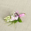 A small yet exquisitely composed boutonnière lies against a neutral woven cloth, its textures and tones working together like a gentle melody. At its core, creamy freesia buds sit poised to open, their petals crisp, almost translucent, and edged with a hint of softness that suggests they were freshly picked that morning. Around them, pale blush-pink flowers unfurl with an easy grace, each petal curving outward in a way that feels like a quiet sigh of contentment. Fine sprays of baby's breath, their tiny white flowers suspended on slender stems, bring a cloudlike airiness to the design, filling the spaces between the blooms without overwhelming them. Long, blade-like strands of greenery weave in and out, giving the boutonnière a subtle sense of movement, while broader leaves, carefully curled and polished, provide a structured, verdant backdrop. The stems are neatly bound with a satiny pink ribbon, wrapped snugly and finished with a tidy knot that suggests both care and affection. The imagined fragrance is gentle but evocative: a sweet, clean aroma that recalls evening walks through Epping's quiet residential streets after a celebration, or a calm moment outside St John's Church before the ceremony begins. The soft, natural lighting across the fabric highlights each curve and contour, from the silky petals to the smooth sheen of the ribbon. Crafted as a finishing touch for a groom, best man, or prom guest, this boutonnière feels personal and intimate, adding a loving brush of colour and nature's quiet artistry to any special day in and around Epping, with the calm assurance of expert local floristry.