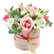 A chic pink hatbox filled with romantic blooms is photographed against a pure white background, creating a clean, modern canvas that lets every detail of the arrangement shine. The cylindrical box is a soft, light pastel pink with a smooth matte texture, gathered at its base with a slim coral-red satin ribbon tied into a neat bow. From this bow dangles a small, cream-coloured gift tag inscribed in elegant green script, suggesting a bespoke floral delivery rather than an off-the-shelf bunch. Rising in a lush, rounded dome above the rim, the flowers appear freshly arranged, as if just set down by a local florist after a walk through Epping High Street. A prominent pale pink rose, its centre swirled with richer pink tones, forms the heart of the design, its velvety petals unfolding in generous layers. Around it, crisp white chrysanthemum blooms with bright green centres add a clear, cheerful contrast, like little bursts of daylight. White alstroemeria weave gently between them, their trumpet-shaped petals marked with fine speckles that introduce quiet intricacy. Scattered through the pastels, deeper fuchsia and hot pink blossoms-probably spray roses or lisianthus-bring lively colour that keeps the bouquet from feeling too reserved. The arrangement is completed with an abundance of greenery: rounded silver-green eucalyptus leaves, deeper green foliage, and slender stems tipped with tiny buds that give a sense of natural growth and movement. The overall effect is elegant yet inviting, a sophisticated hatbox bouquet perfectly suited to birthdays, anniversaries, new baby celebrations, or simply sending love across Epping and the nearby villages with a gift that feels personal and beautifully considered.