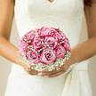This photograph focuses closely on a bridal bouquet of blush-pink roses and white baby's breath, cradled against an elegant lace wedding dress. The roses are gathered into a compact, almost perfectly spherical shape, each bloom at its peak with petals that unfurl in smooth, satiny layers from the centre. Their colour is a gentle gradient of pale blush through to a soft, romantic pink, mirroring the delicate hues often seen around Epping's historic churches on bright spring mornings. Around the roses, a mist of white gypsophila softens the outline of the bouquet, creating a feathery, cloud-like border that blends seamlessly into the bride's ivory lace bodice. The lace itself is detailed and intricate, with floral motifs that echo the shapes of the rose petals, adding depth and texture behind the bouquet. Daylight illuminates the scene, likely streaming in from a nearby window of a country venue near Epping Forest, highlighting the subtle gloss on each petal and the tiny green stems of the gypsophila. The bouquet rests in the bride's hands with a sense of ease and quiet confidence, suggesting a moment of stillness before the ceremony begins at a local registry office or nearby village church. You can almost sense the coolness of the rose petals against the fingertips and imagine the soft, sweet fragrance that would drift up as she walks. Designed with the thoughtful care of a local Epping florist, the arrangement feels timeless and deeply personal, a gentle expression of love and hope that will carry her from the first step down the aisle to the final dance of the evening.