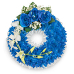 A formal circular sympathy wreath composed almost entirely of densely packed, electric royal blue chrysanthemums - their small, closely cupped petals creating a plush, velvet-like ring that reads both vivid and solemn against a bright white backdrop. At the top centre, a trio of deep blue roses opens fully, their velvety petals catching a cool, even light, and they are gently haloed by tiny clusters of pure white baby's breath and a few deep green leaves that punctuate the blue with natural contrast. From the upper left quadrant a graceful spray of pristine white orchids (likely Phalaenopsis or Dendrobium) descends, each blossom with broad, delicate petals arranged along a slender, arching stem; interspersed among them are soft pale yellow-green buds that suggest quiet renewal. The wreath's inner edge is neatly finished with a ruffled white satin ribbon that frames the central void and lends a crisp, attentive finish to the composition. The whole arrangement sits in neutral light that emphasises texture - the chrysanthemums' dense mass, the roses' smooth richness, and the orchids' pearly translucence - and it carries a faint, clean floral scent reminiscent of a chapel on a calm morning. Intended as a dignified tribute for a funeral or memorial, this piece balances bold colour with gentle white accents to convey serenity, remembrance and respect. Crafted with local care in Epping - close to the Market Square and Epping Forest - the wreath is a considered choice for families seeking a composed, reverent floral expression at services held near St. John's Church or at local funeral homes.