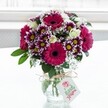 A vibrant mixed bouquet of deep magenta gerbera daisies, purple-tipped chrysanthemums, and creamy white roses stands proudly in a clear, textured glass vase, centred against a bright, softly diffused white background that feels like daylight streaming through an Epping kitchen window. The gerberas, with their velvety fuchsia petals and dark, almost inky centres, draw the eye first, radiating cheerful energy. Around them, clusters of daisy-like chrysanthemums display intricate petals that are rich purple at the tips and gradually fade to white near their sunny yellow centres, adding both movement and depth. Nestled between these bolder blooms are delicate white spray roses, some fully open, others still in tight bud, lending a gentle, romantic softness. Wisps of airy white baby's breath weave through the arrangement like tiny stars, lightening the palette and giving the bouquet a whimsical texture. Lush deep green foliage frames the flowers and provides a sturdy base, the leaves catching the light with a quiet sheen. The squat glass vase is cinched at the neck with a simple rustic twine bow, and from it hangs a small white rectangular gift tag printed with a pink rose emblem and elegant script, suggesting a thoughtful, locally delivered gift rather than a supermarket bunch. The overall effect is fresh, joyful, and polished, the kind of arrangement you might have sent from a trusted Epping florist for a birthday on the High Street, a congratulations bouquet in Theydon Bois, or a pick-me-up delivered to a home overlooking Epping Forest, bringing colour, texture, and a sense of celebration into any room.