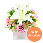 This image shows a refined pink and white floral centerpiece standing crisply against a pure white background, reminiscent of a freshly arranged bouquet ready to be sent from a local Epping florist. At the centre, two fully open white Oriental lilies spread their broad, smooth petals with effortless grace, their interiors marked by strong reddish-brown stamens that lend depth and structure. Nestled closely around these lilies are numerous soft pink roses, some tightly wound and others generously opened, revealing velvety whorls of petals that feel tender and romantic. Among the roses and lilies, delicate stems of pink and white alstroemeria introduce a hint of playful pattern with their slender petals and soft yellow speckling. Tiny sprays of white baby's breath appear throughout the arrangement like little puffs of cloud, adding lightness and gently filling in any gaps. A lush assortment of greenery-broad, glossy leaves, slim grass-like blades, and fine, feathery fronds-surrounds and supports the blooms, evoking the natural abundance of gardens near Epping Forest and the leafy streets leading toward Theydon Bois. The flowers are gathered into a clear, square glass vase, the transparent sides revealing clean stems and water, giving the design a neat, contemporary look that suits a dining table, office desk, or cosy corner in a home off Epping High Street. Wrapped around the middle of the vase is a soft, pale pink satin ribbon, adding an understated decorative accent. In the lower right corner of the image, a bright circular orange emblem with bold white capital text reads VASE INCLUDED, immediately clarifying that the container comes with the arrangement. The overall feel is elegant, fresh, and quietly celebratory, ideal for thoughtful deliveries across Epping.